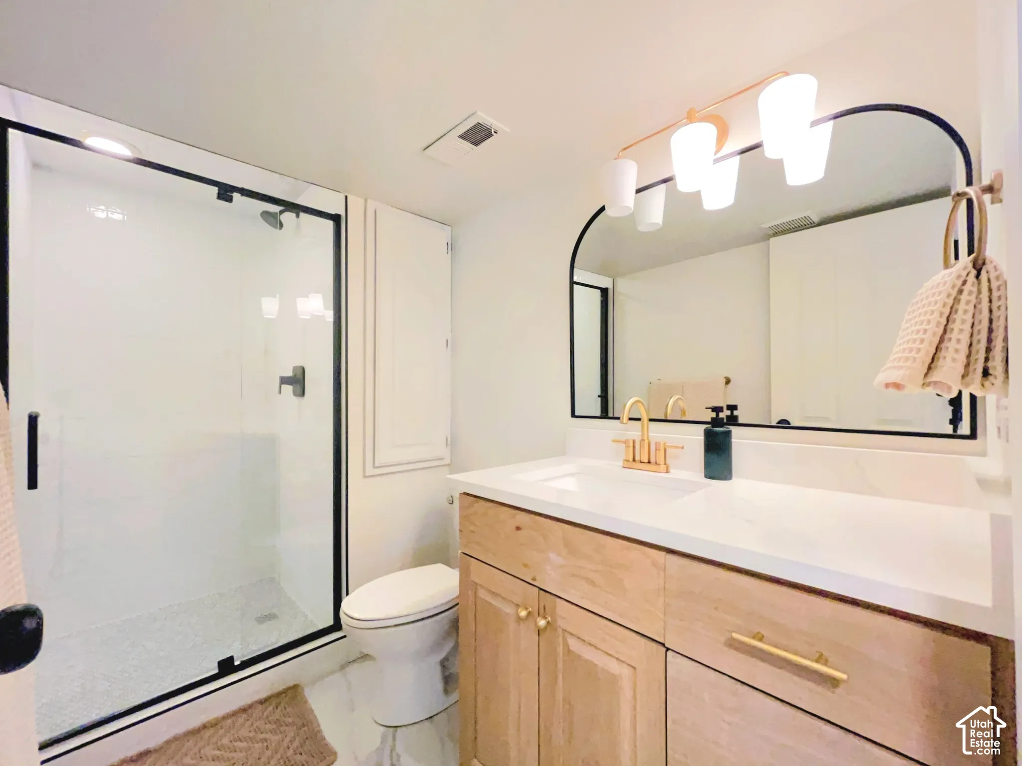Frameless glass shower enclosure with matte black trim, natural wood-finish vanity with gold-tone hardware, arched mirror, contemporary vanity light fixture, and white countertop