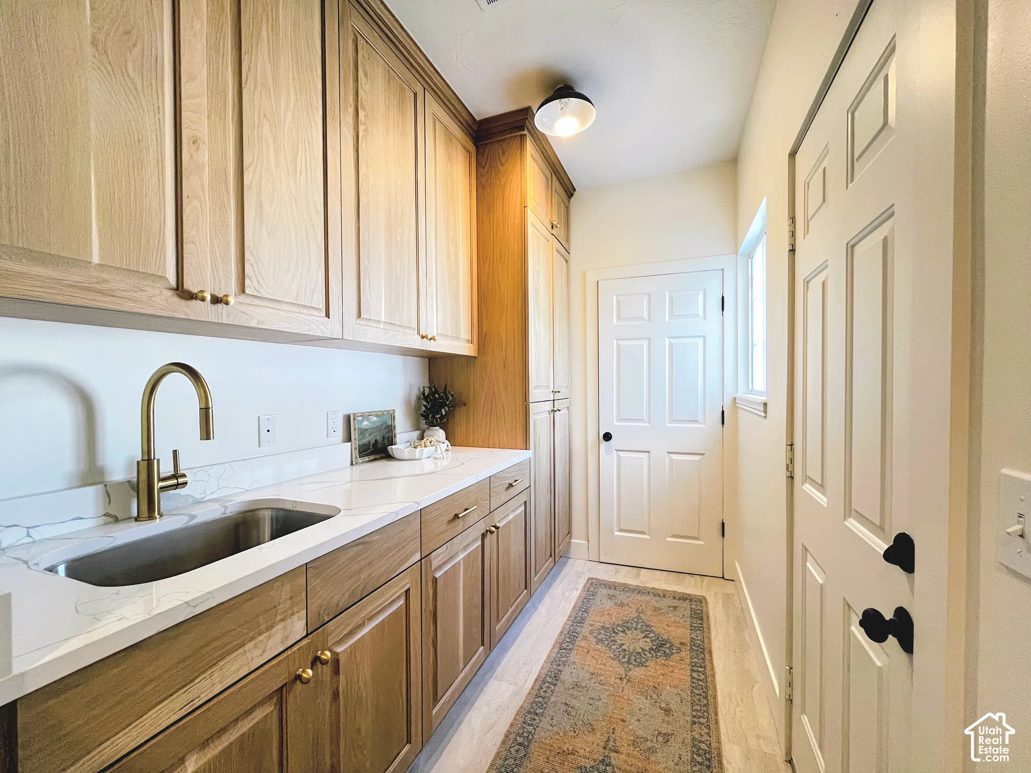 White solid surface countertop with integrated undermount sink, brass finish pull-down faucet, light wood-finish cabinetry, and light wood-finish flooring