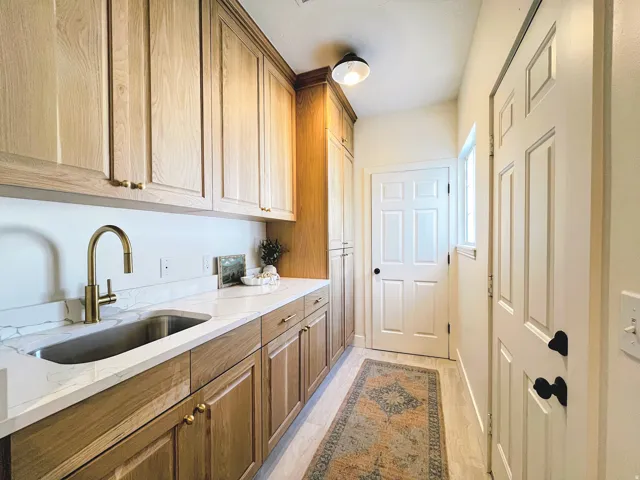 White solid surface countertop with integrated undermount sink, brass finish pull-down faucet, light wood-finish cabinetry, and light wood-finish flooring