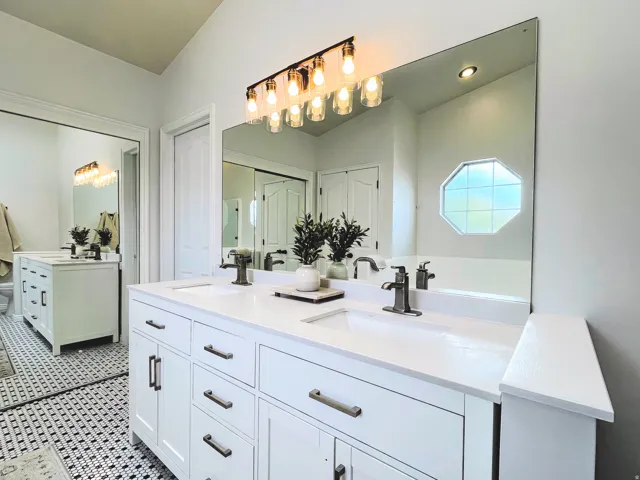 Dual white vanities with integrated sinks, brushed nickel fixtures, a large frameless mirror, and a black and white mosaic tile floor