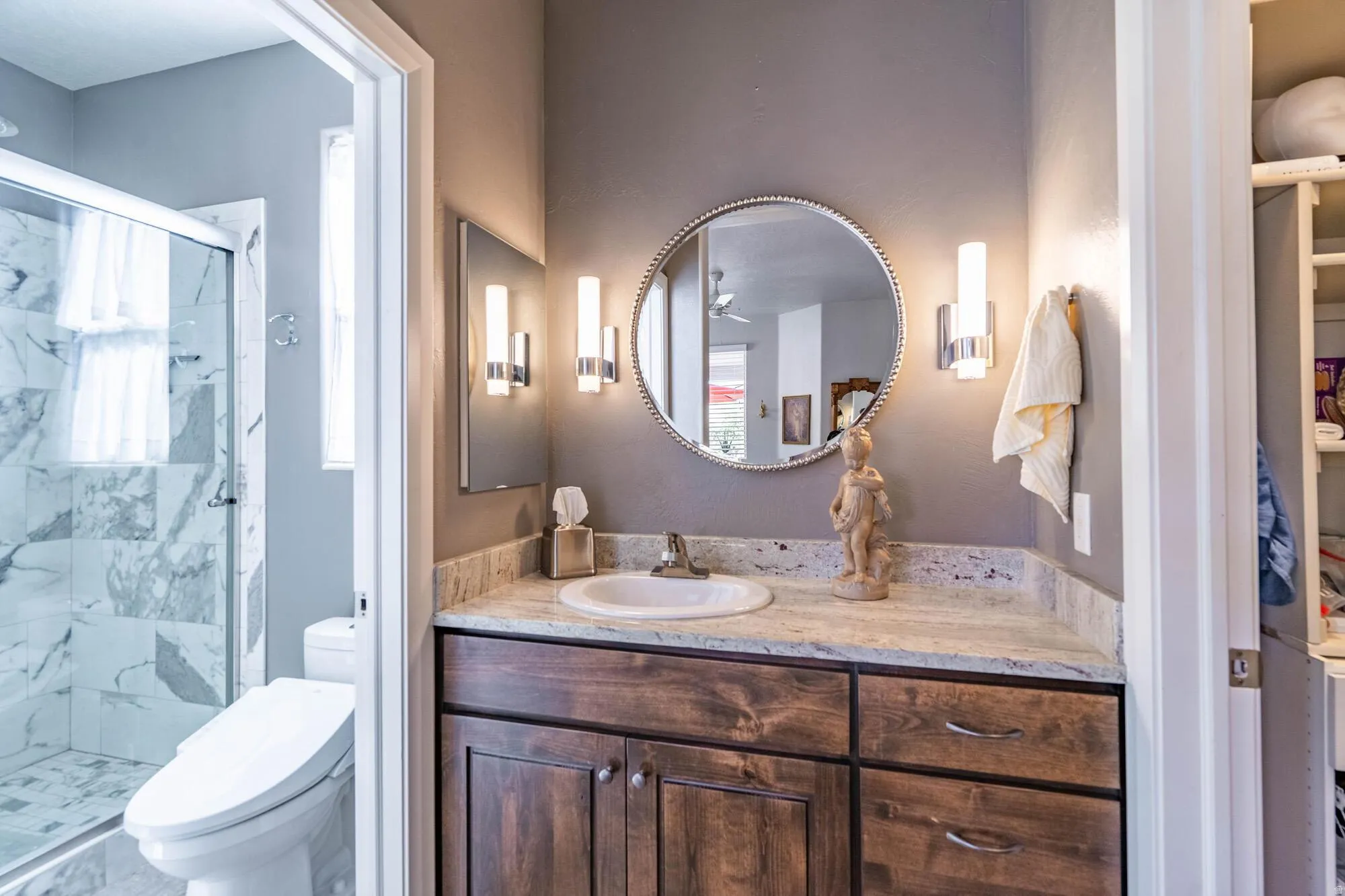 Granite-look countertop with an undermount sink, wood-finish vanity with cabinet storage, and a framed circular mirror