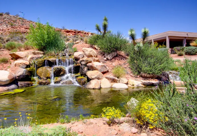Rock waterfall feature with a pond, surrounded by desert landscaping and mature shrubs, with a building structure in the background