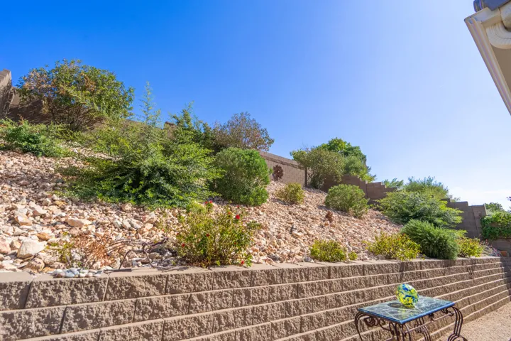 Multi-tiered retaining wall system, mature landscaping, rock-filled hillside, and clear sky