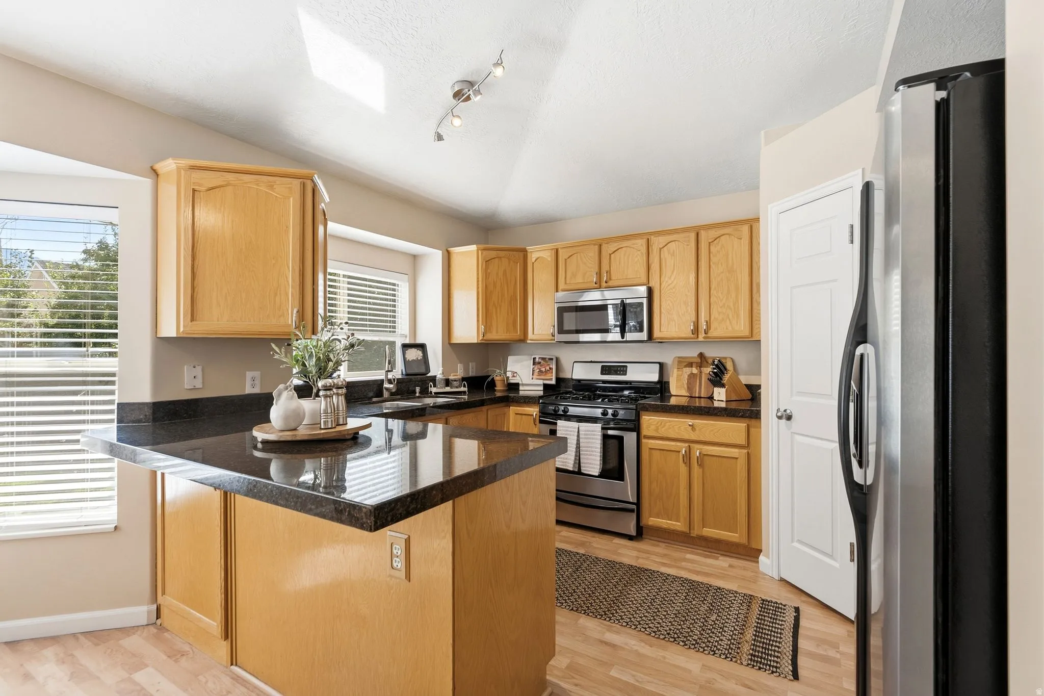 Kitchen featuring light wood cabinetry, dark countertops, stainless steel appliances, and wood-finish flooring