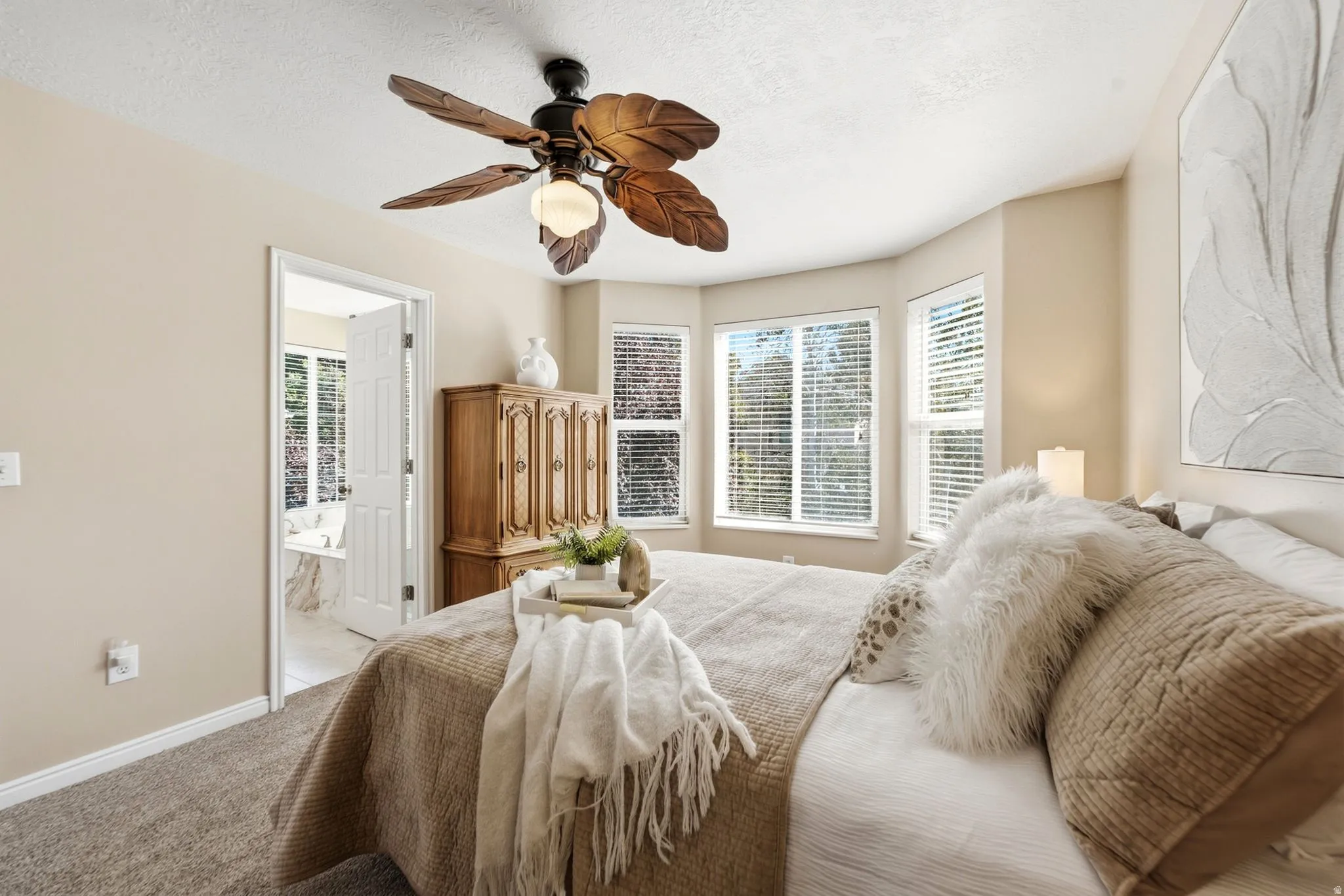 Bay window with multiple windows, ceiling fan with decorative blades, wall-to-wall carpeting, neutral wall color, and an open doorway