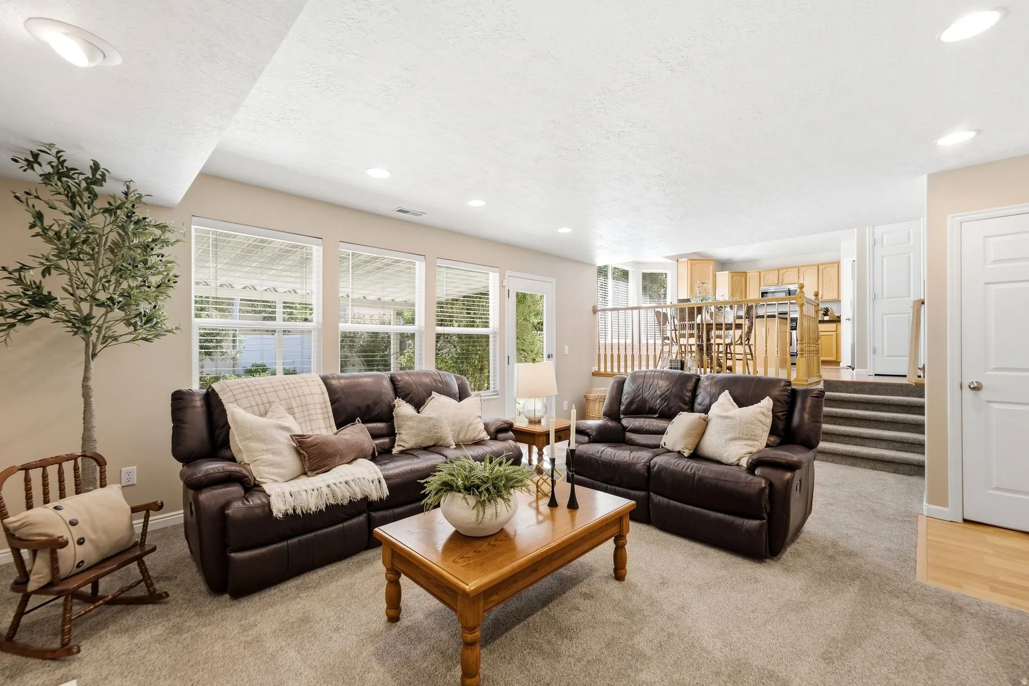 Recessed lighting, light beige wall paint, multiple windows with blinds, light wood-finish flooring, and a partial view of a kitchen with light wood cabinetry