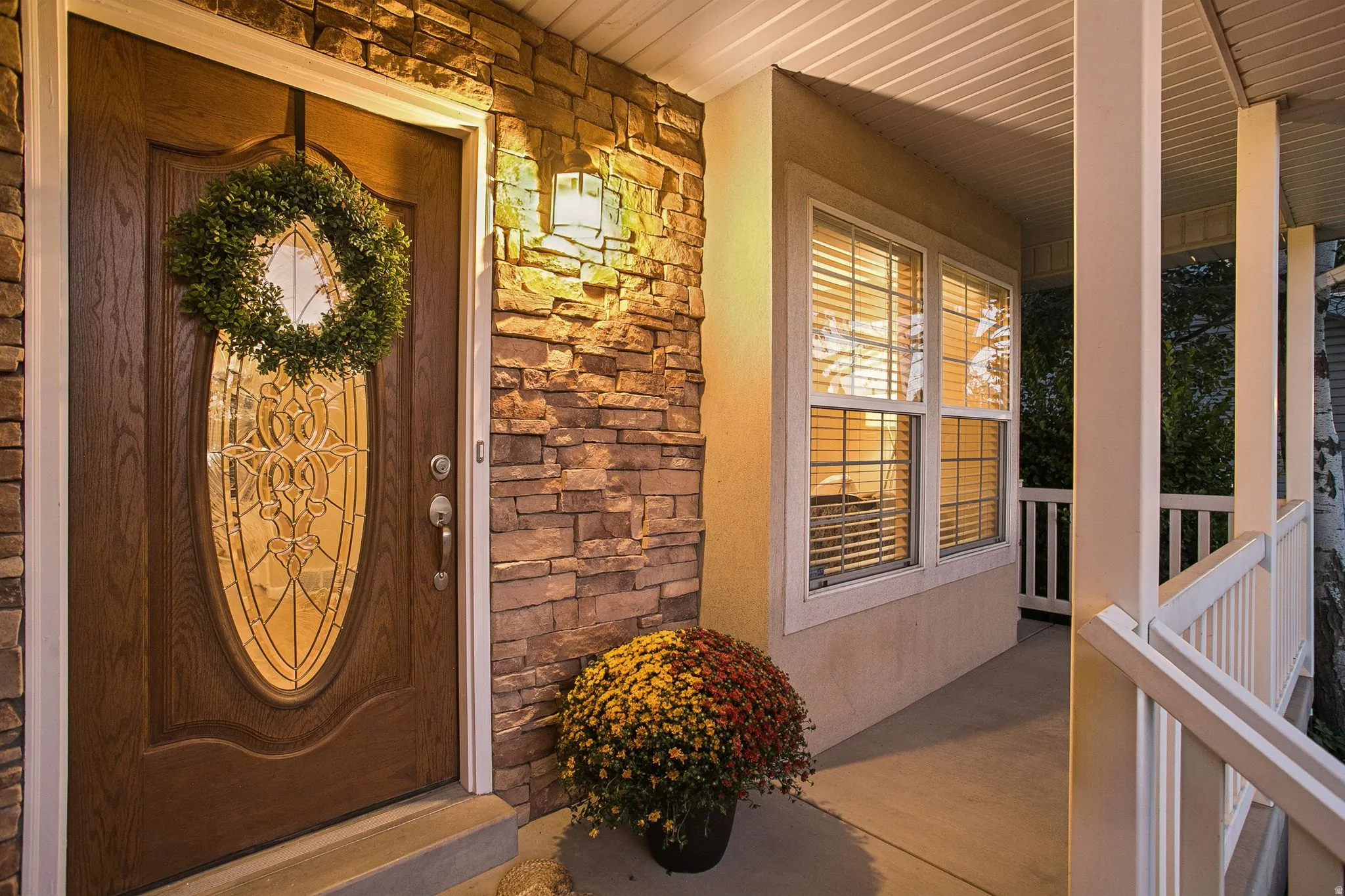 Wood-finish entry door with decorative glass insert, stone veneer exterior accents, covered porch with white railing, and double-hung windows with blinds