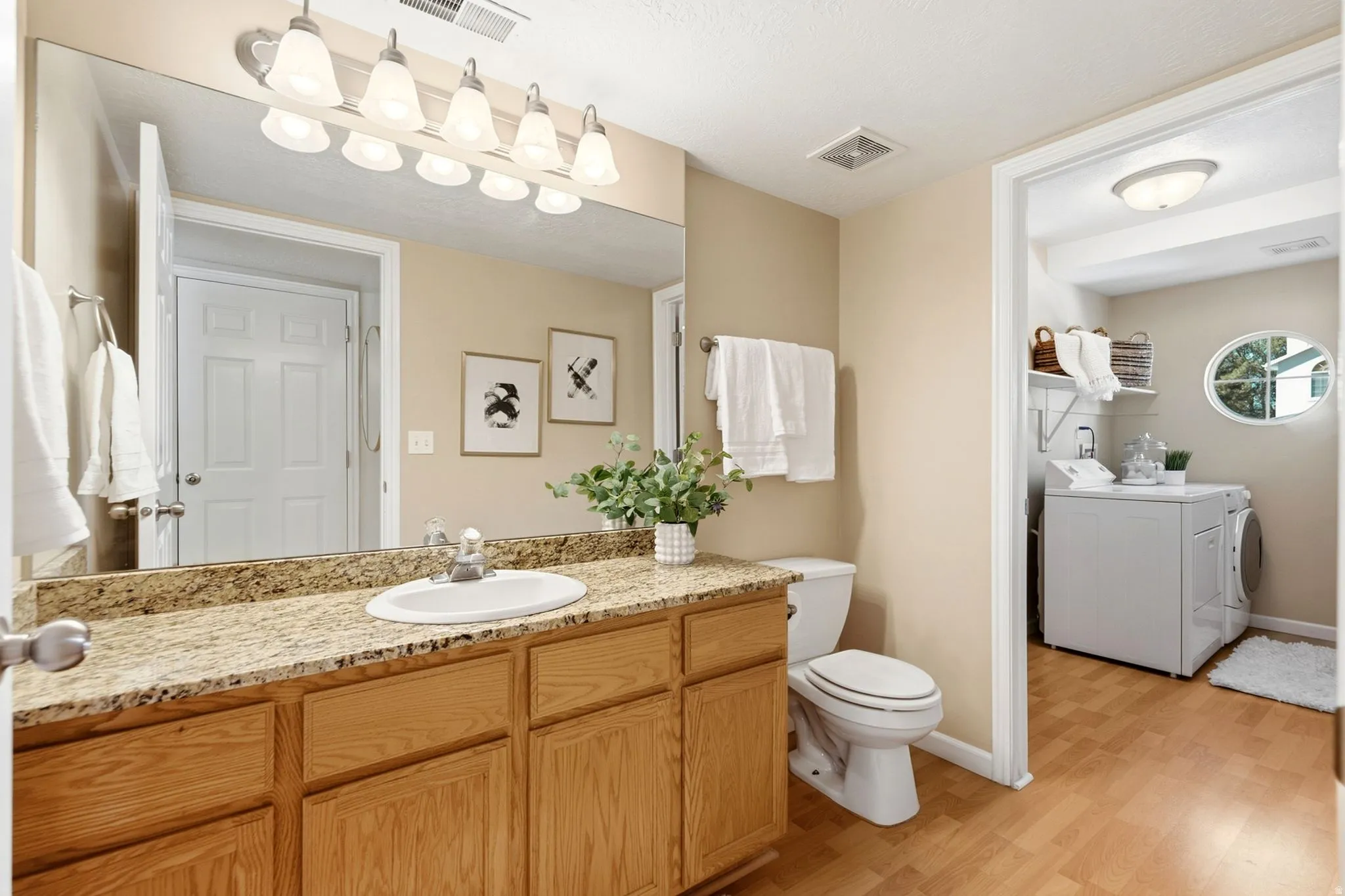 Wood-finish flooring, vanity with granite countertop and integrated sink, full-width mirror, and adjacent laundry area with a round window