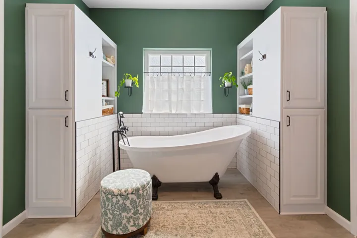 Freestanding clawfoot bathtub with wall-mounted fixture, framed window, full-height built-in cabinetry, and subway tile wainscoting