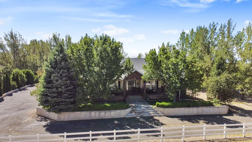Expansive paved driveway, mature trees, concrete retaining wall with landscaping, and white fencing