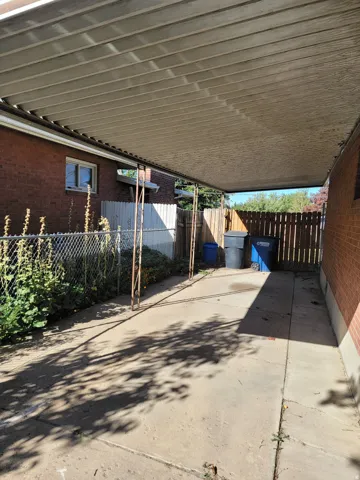 Covered patio area with concrete surfacing, supported by ornate metal posts, bordered by a chain-link fence and a wood fence, alongside a brick exterior wall