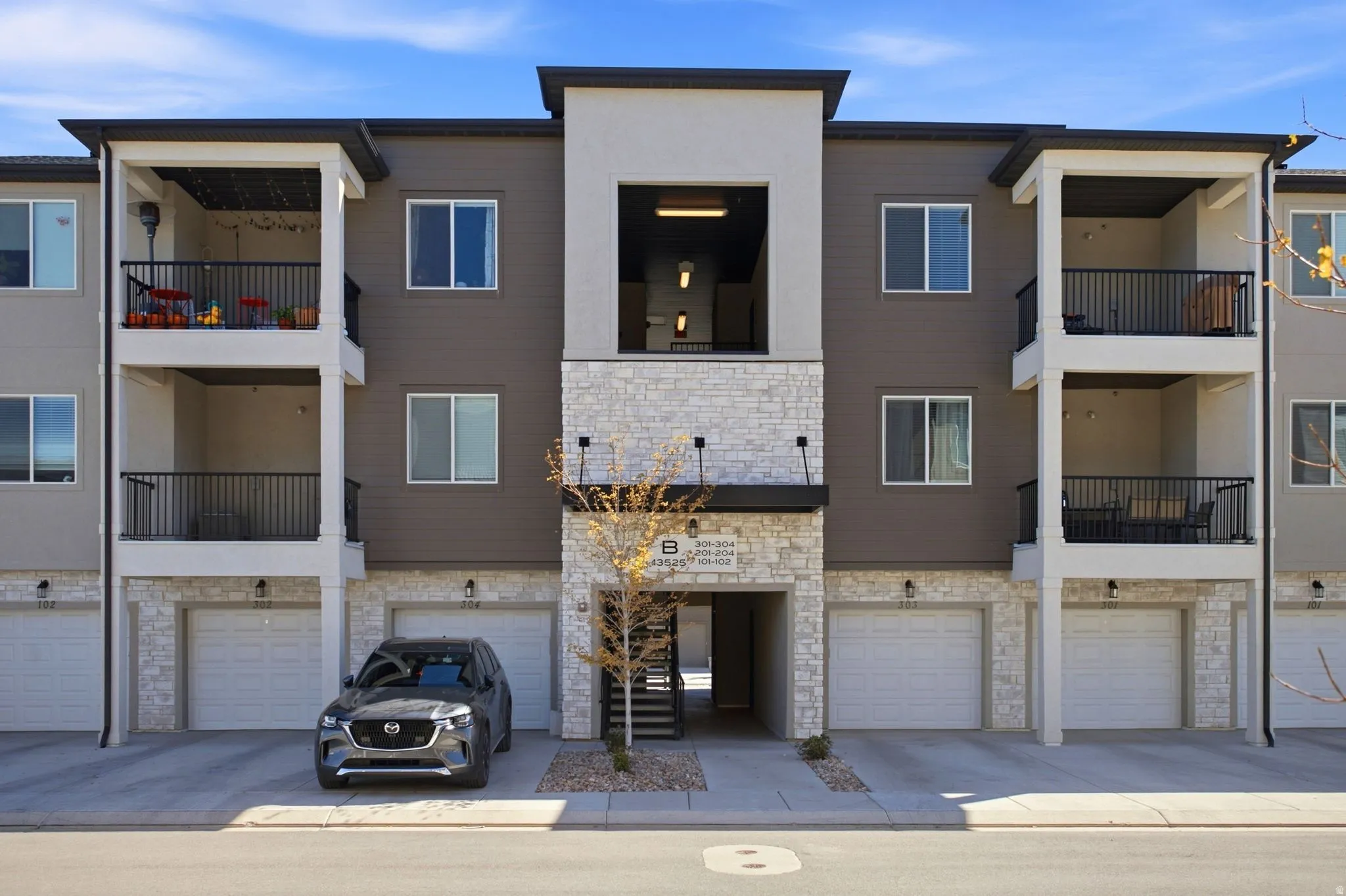 Multi-story building featuring stacked balconies with black railings, individual ground-level garages, a central stone-clad entrance, and a modern exterior facade with contrasting siding