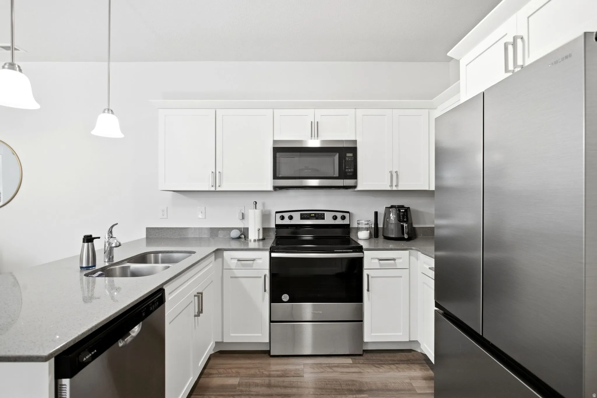 Modern kitchen featuring white shaker-style cabinetry, stainless steel appliances, light-toned countertops, wood-finish flooring, and pendant lighting