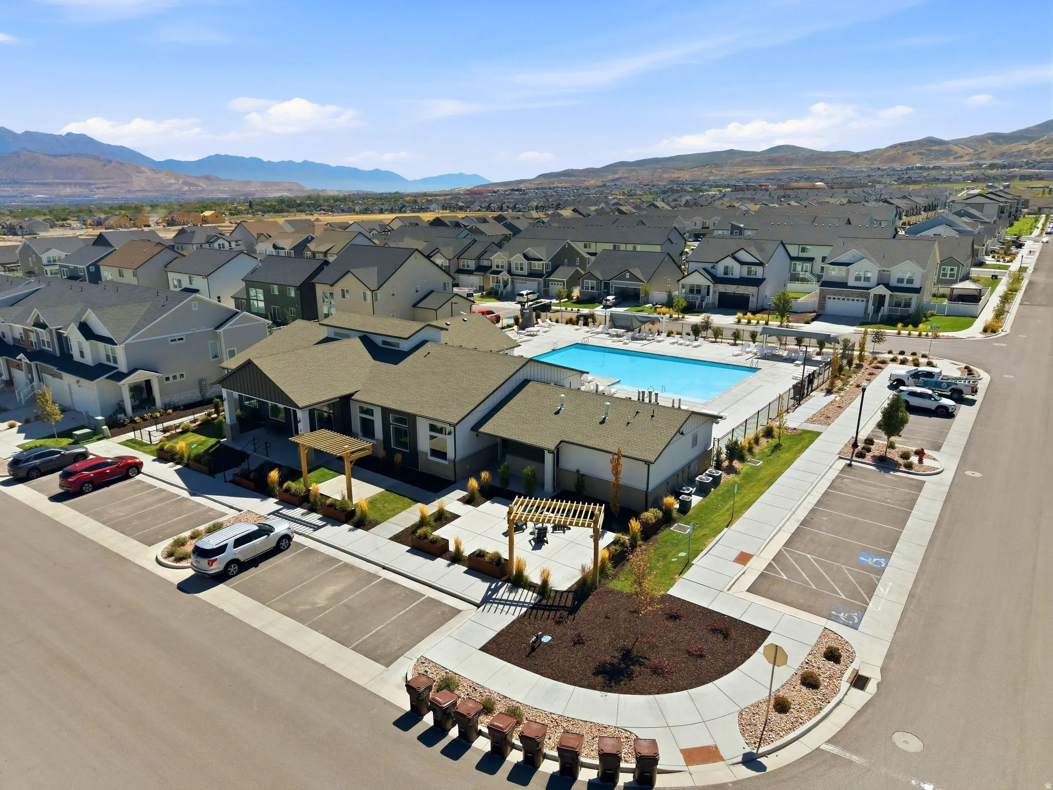 Community pool with surrounding patio, clubhouse with exterior pergola, paved parking, and a manicured landscape