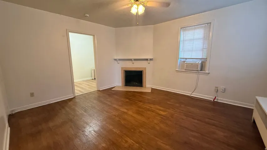 Wood-finish flooring, corner fireplace with stone surround, ceiling fan, and window with blinds