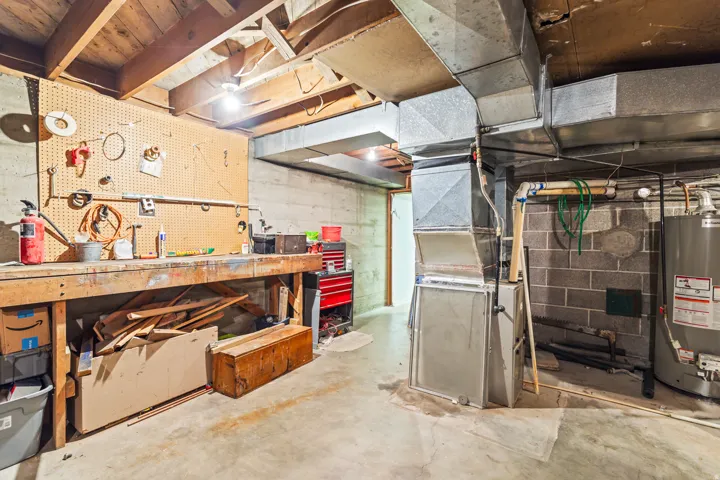 Unfinished basement with exposed ceiling joists, concrete block walls, a dedicated workbench with pegboard, and visible ductwork