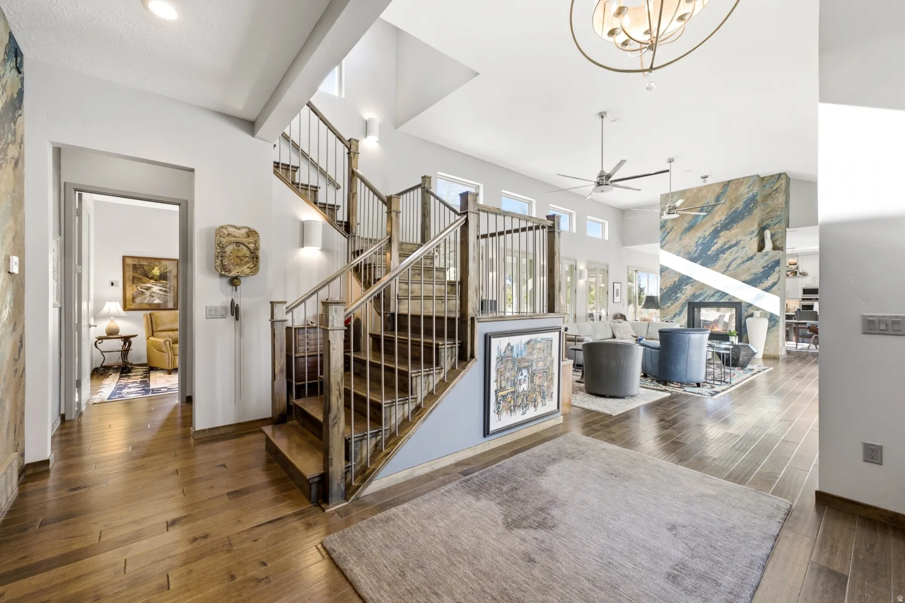 Expansive open-concept living space with wood-finish flooring, a contemporary floating staircase with metal balusters, high ceilings, and a fireplace with a striking accent wall