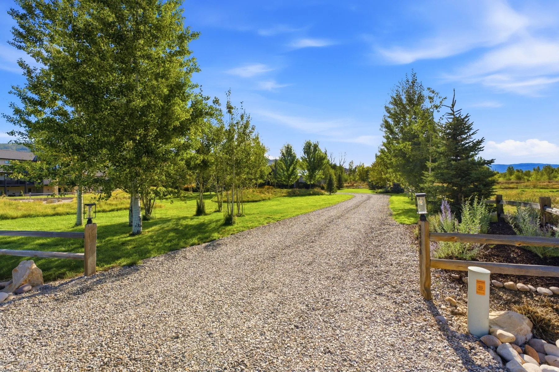 Gravel drive, wood post fencing, mature trees, landscaped grounds, and clear blue sky