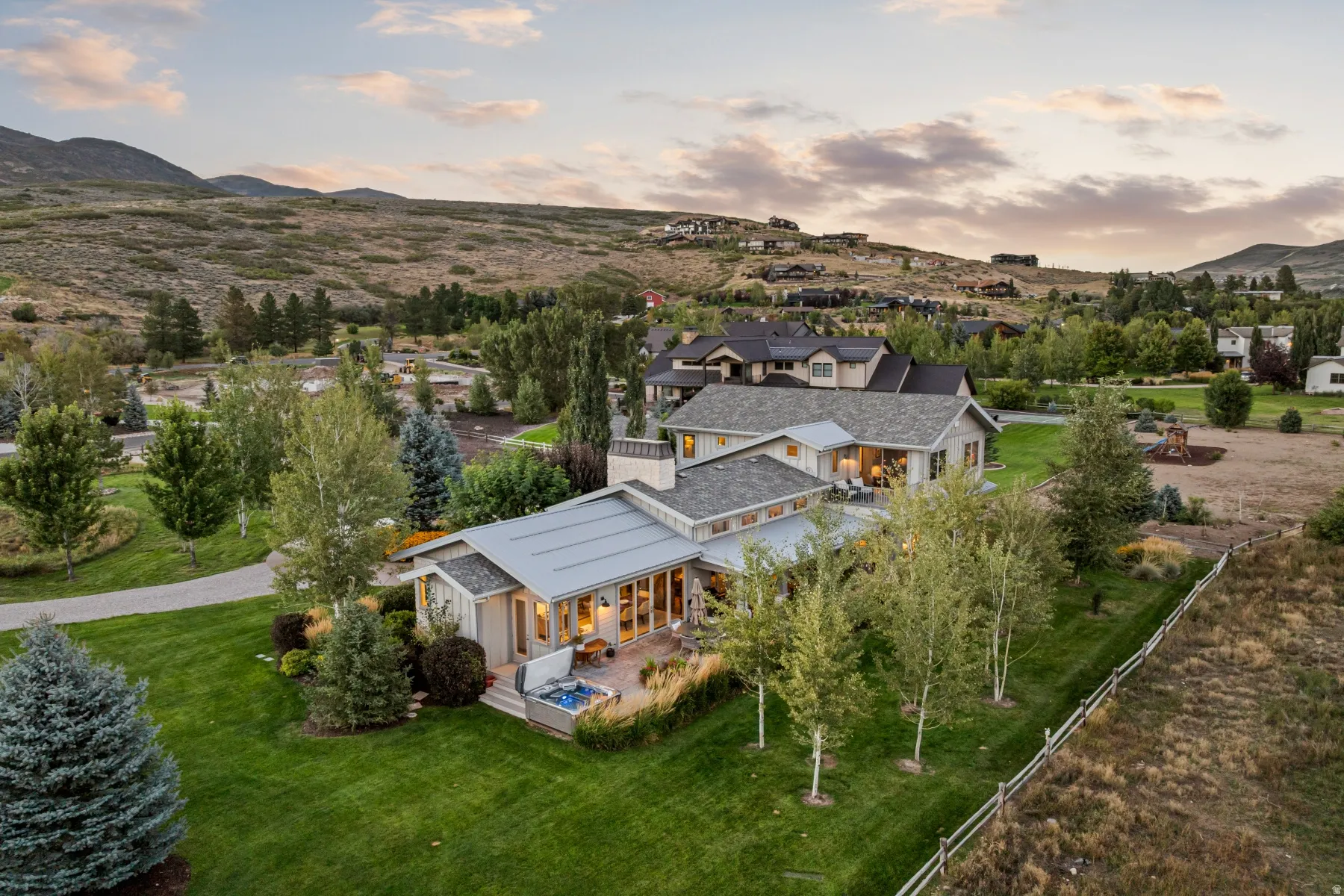 Expansive estate featuring a stucco and siding exterior, multiple gabled roofs, a paved patio with a hot tub, a large manicured lawn, and mature trees