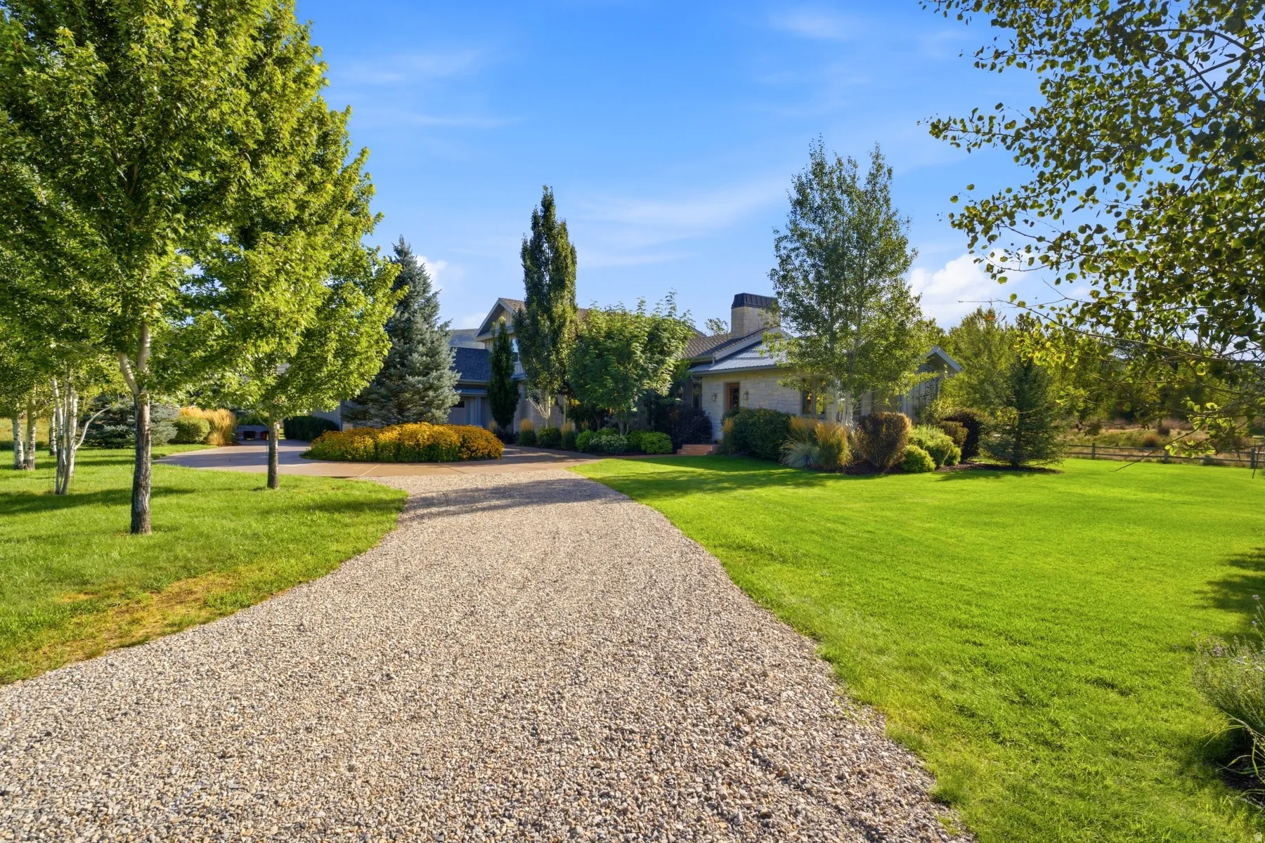Expansive gravel driveway leading to a residence with a blend of stucco and wood siding, featuring a prominent chimney and mature landscaping