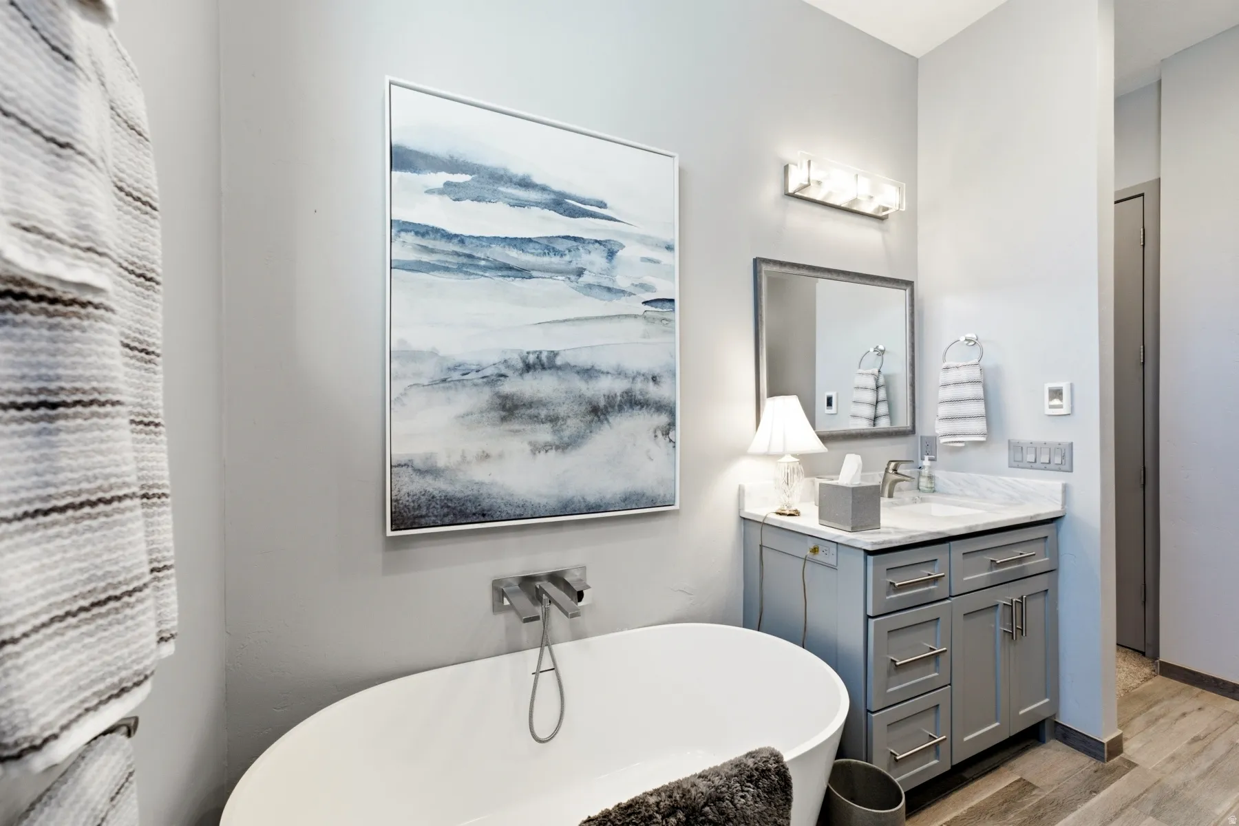 Freestanding soaking tub with wall-mounted faucet, single vanity with light-gray cabinetry, and stone countertop