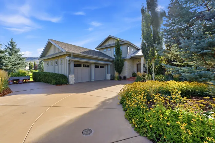 Paved driveway leading to a three-car garage, stone and siding exterior, mature evergreen trees, and vibrant yellow landscaping