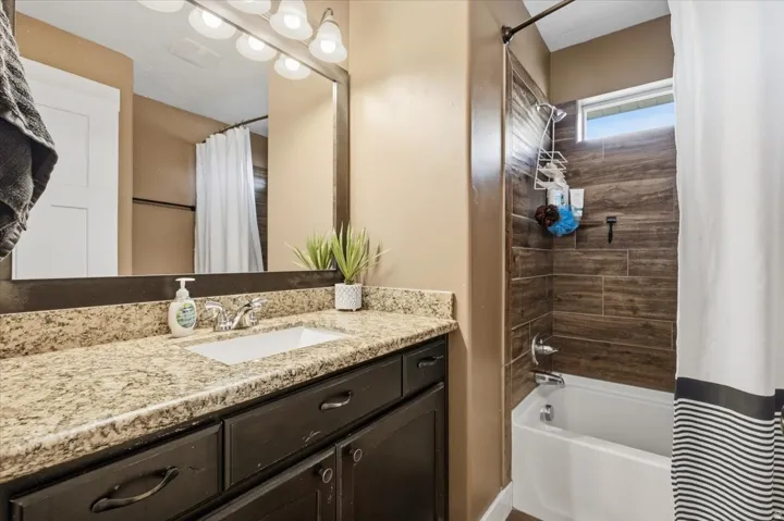 Bathroom featuring a granite-look countertop, dark wood-finish vanity, undermount sink with chrome-finish faucet, mirror with overhead lighting, and a bathtub with wood-look tile surround