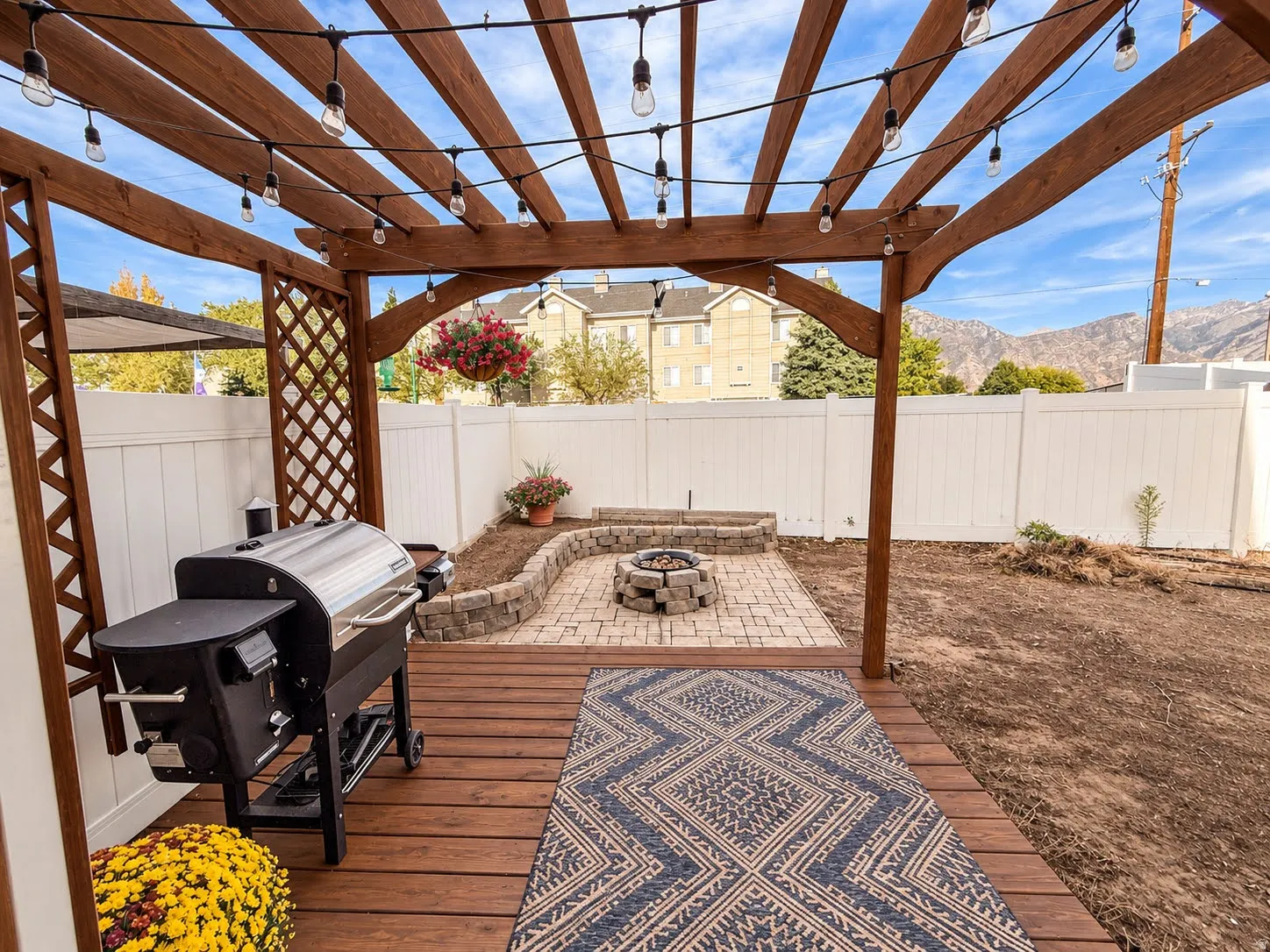 Wood pergola with string lighting, wood deck, paver patio, stone fire pit, and white privacy fencing