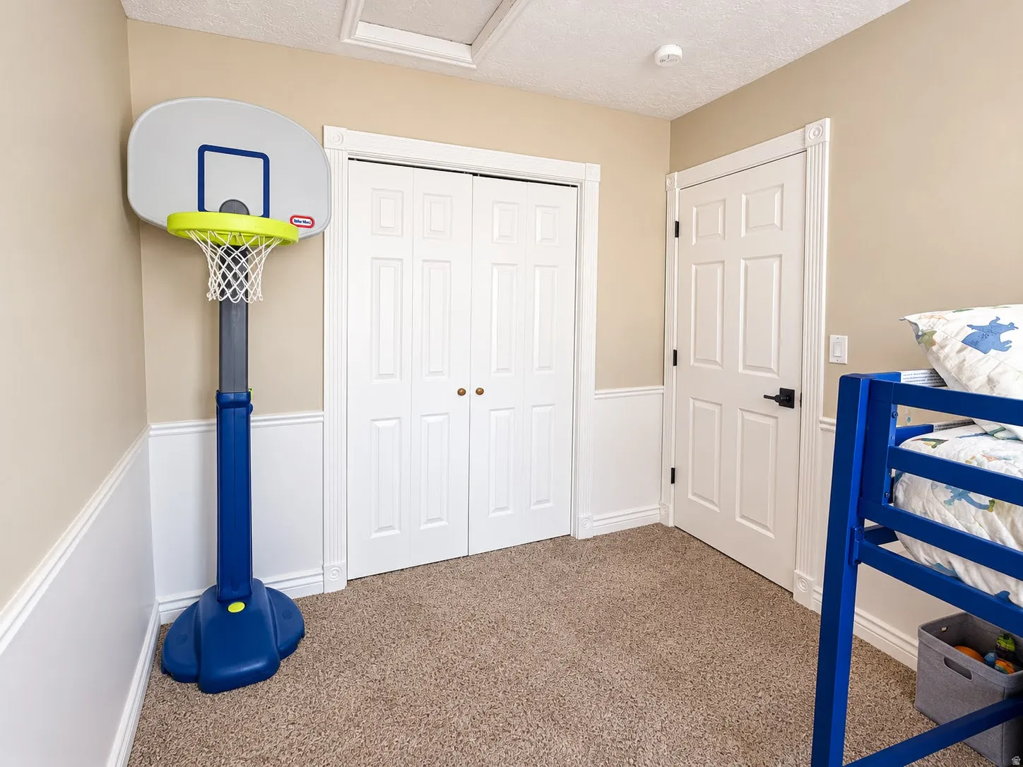 Neutral tone wall paint, white wainscoting, white bi-fold closet doors, textured carpet flooring, white paneled interior door with matte black hardware