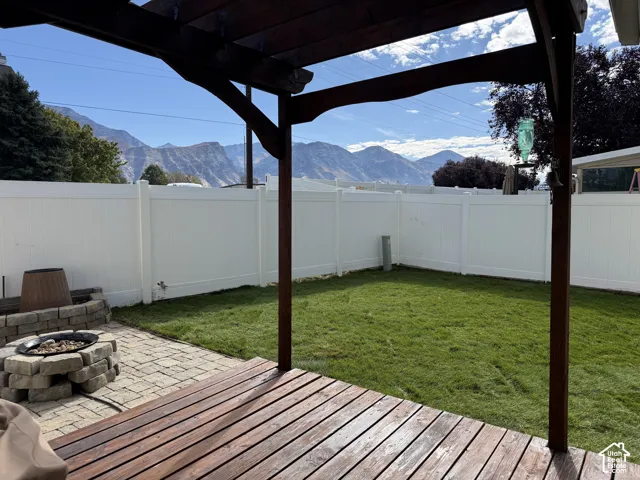Wooden deck with an overhead pergola, paver patio featuring a stone-look fire pit, green lawn, and white privacy fencing