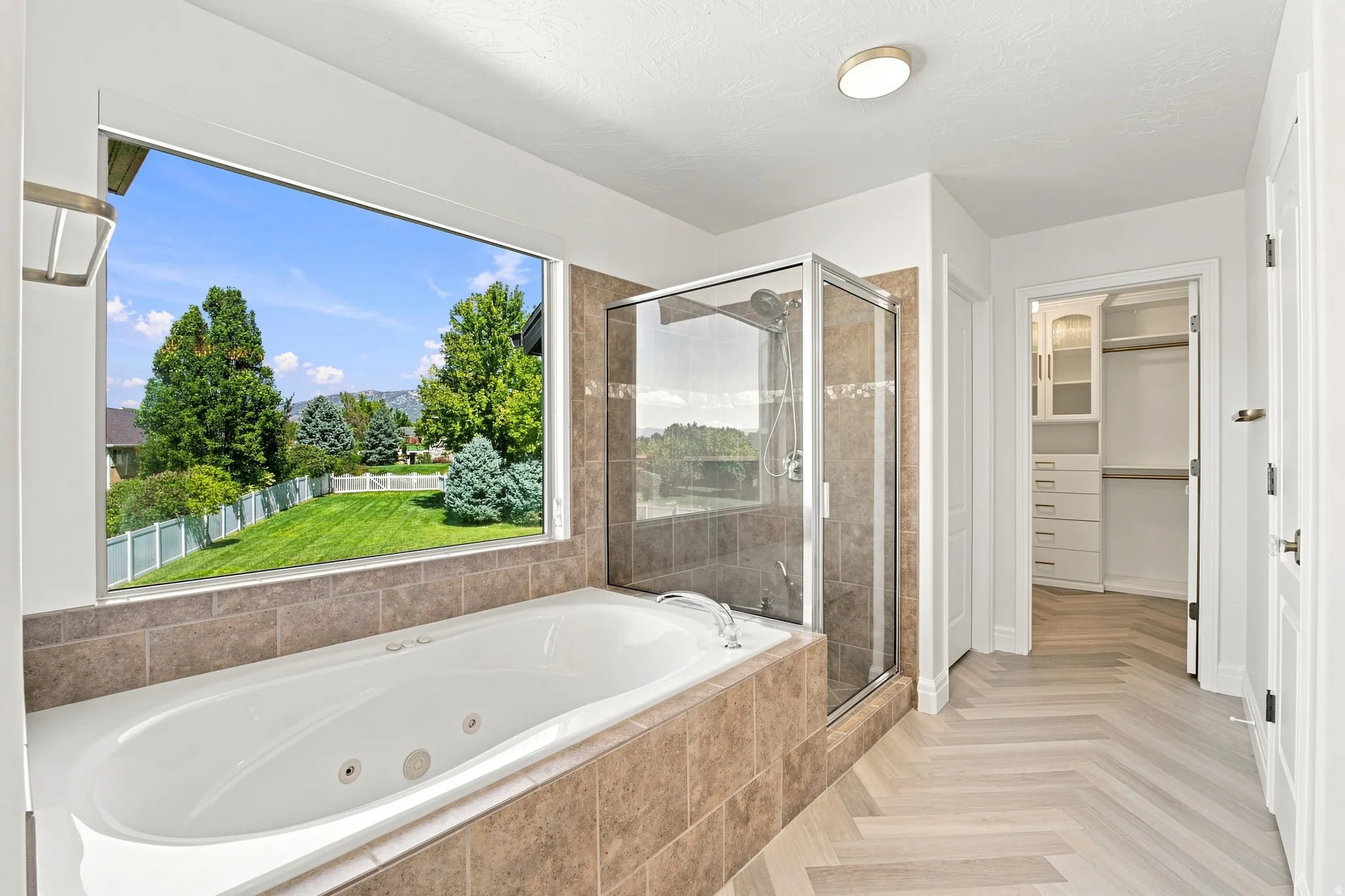 Soaking tub with tile surround, glass-enclosed shower, herringbone wood-finish flooring, built-in closet organization, and large picture window