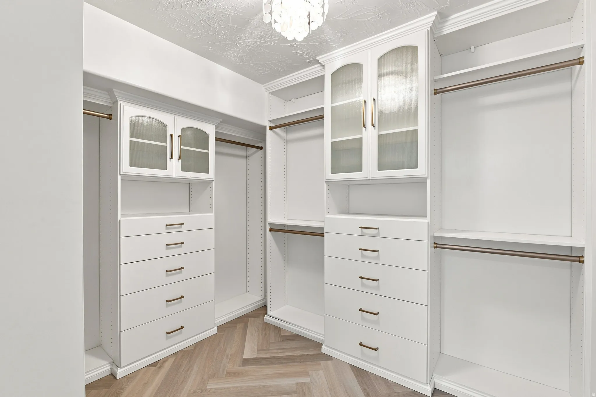 Custom closet system featuring white cabinetry, glass-front upper cabinets, built-in drawers with bronze hardware, and wood-finish herringbone flooring