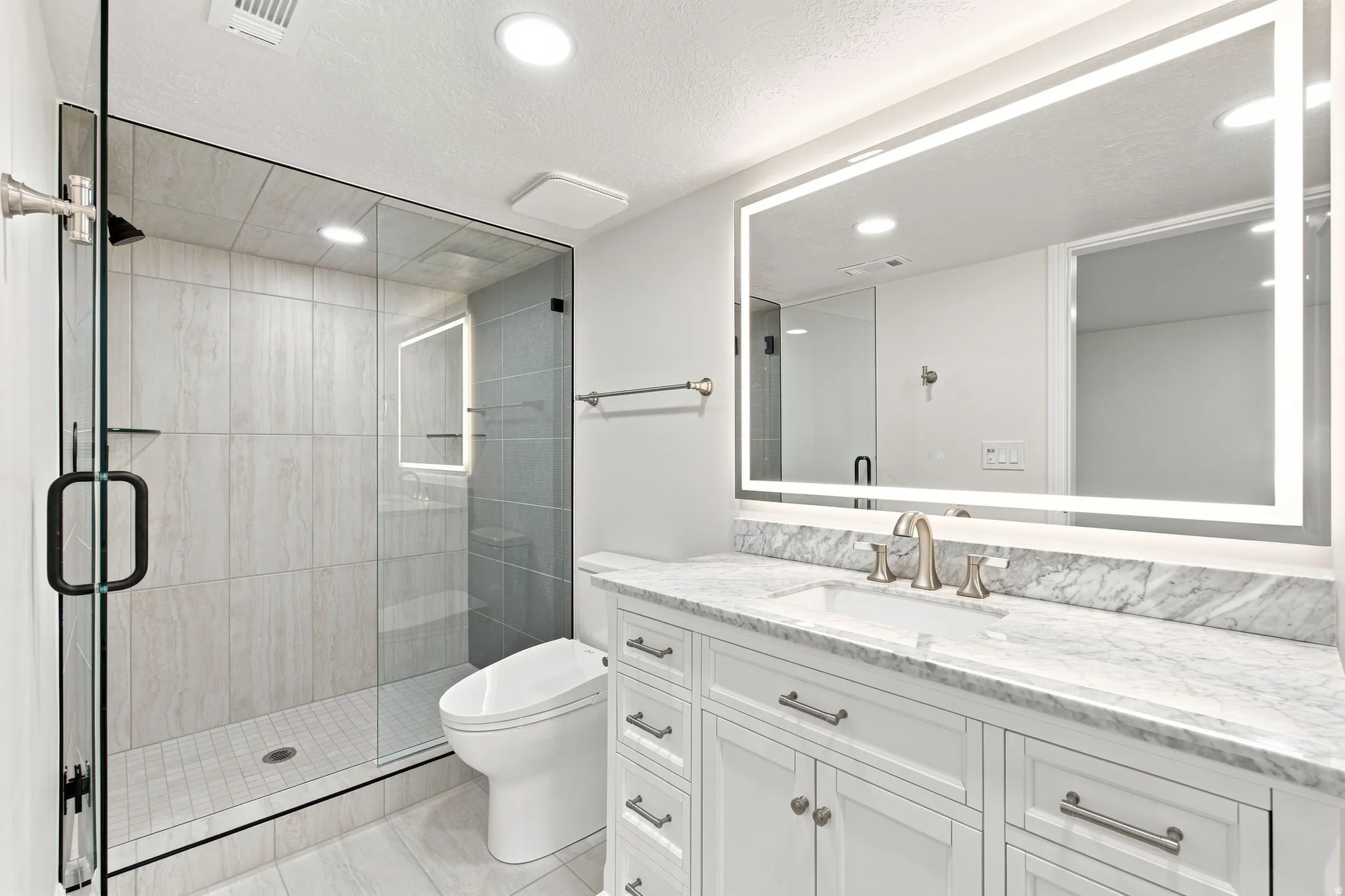 Glass-enclosed shower with subway tile surround, white vanity with stone countertop, undermount sink, and illuminated mirror