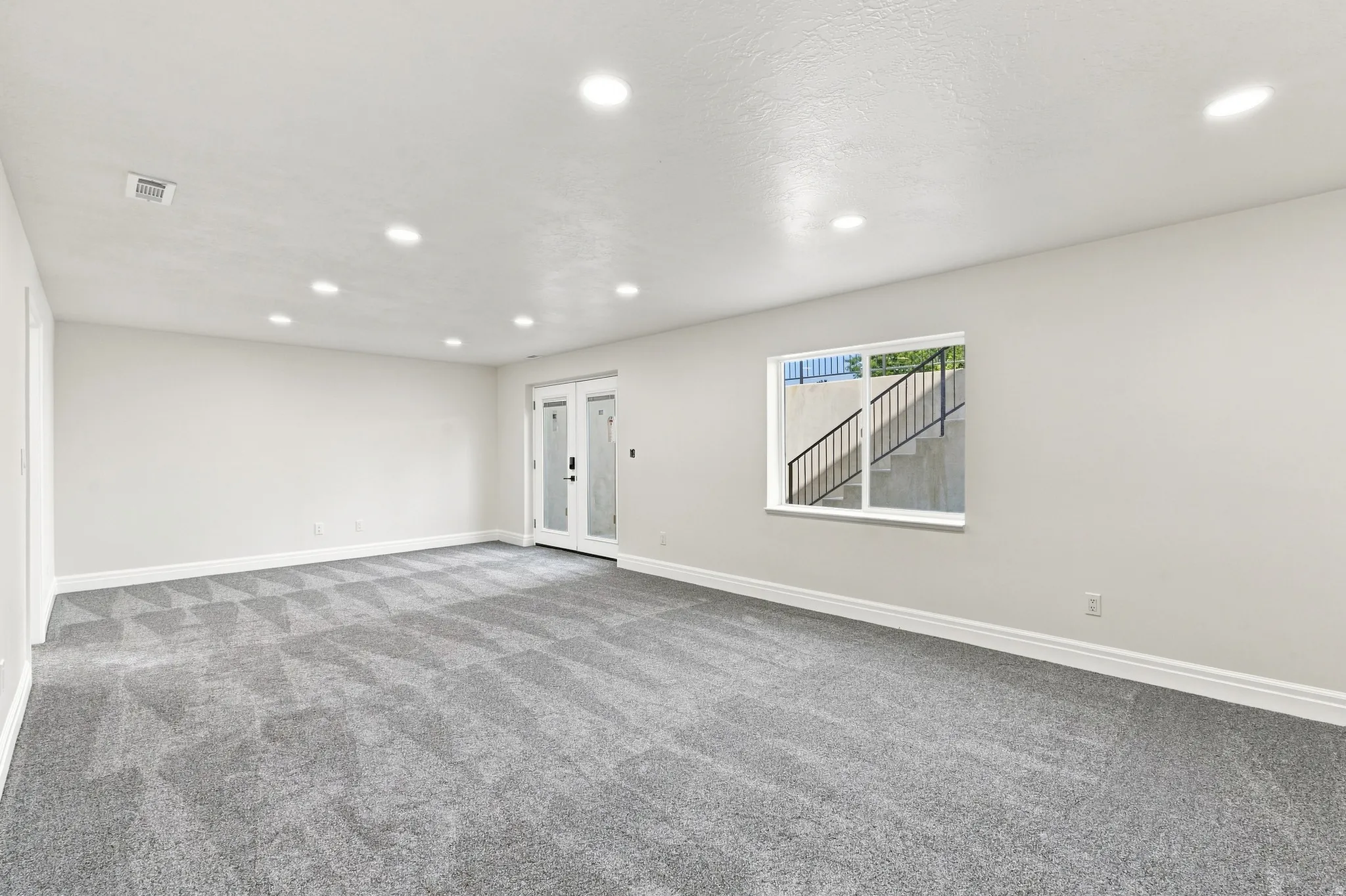 Spacious room featuring gray patterned carpeting, recessed ceiling lighting, a white framed window, and white French doors