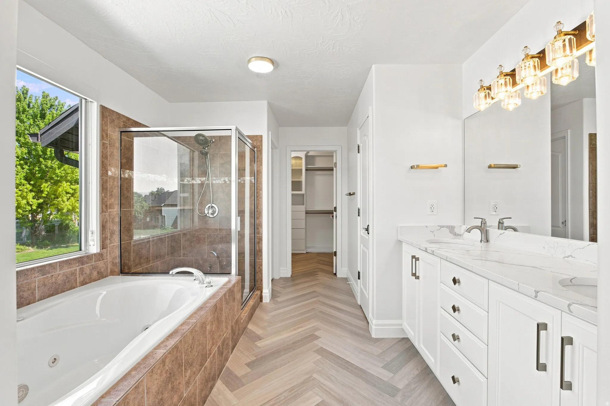 Herringbone wood-finish flooring, dual vanity with undermount sinks and light-toned countertops, built-in soaking tub, glass-enclosed shower, and a walk-in closet with built-in shelving