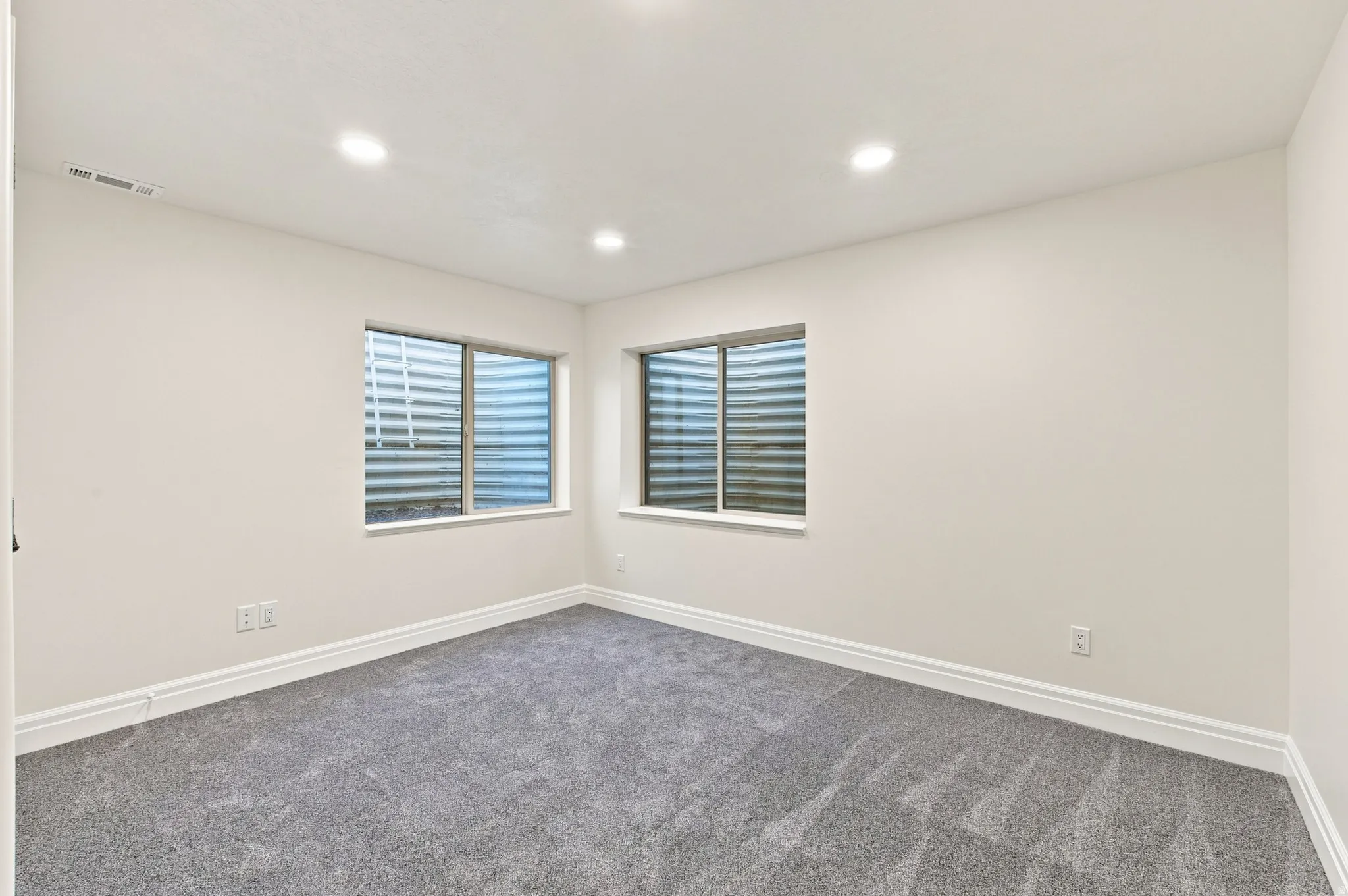 Recessed lighting, light-colored walls, two windows with window wells, gray carpeting, and white baseboards
