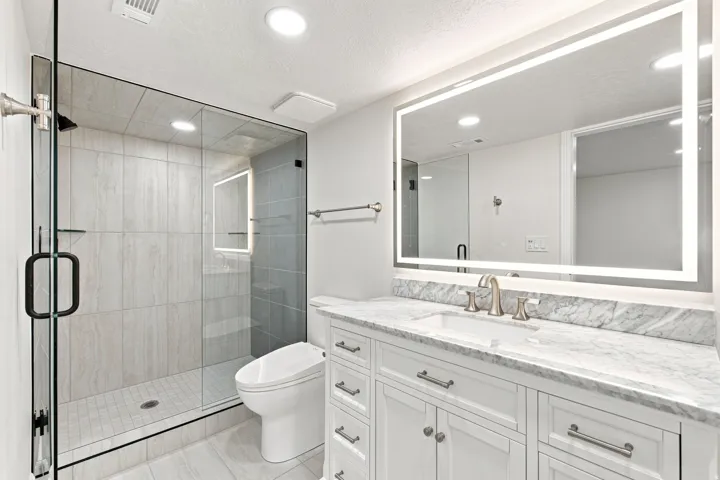 Glass-enclosed shower with subway tile surround, white vanity with stone countertop, undermount sink, and illuminated mirror