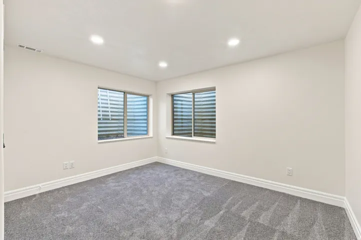 Recessed lighting, light-colored walls, two windows with window wells, gray carpeting, and white baseboards