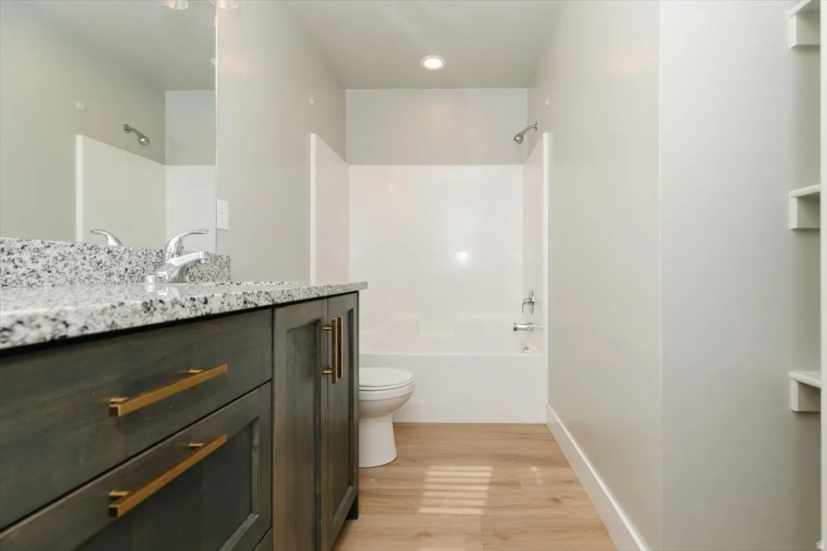 Bathroom featuring a vanity with a speckled countertop, a chrome faucet, a white tub with integrated shower, wood-finish flooring, and built-in shelving