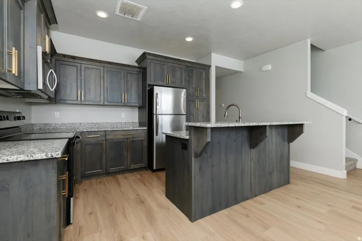 Kitchen featuring wood-finish cabinetry, granite countertops, stainless steel appliances, a kitchen island with an integrated sink, and wood-finish flooring