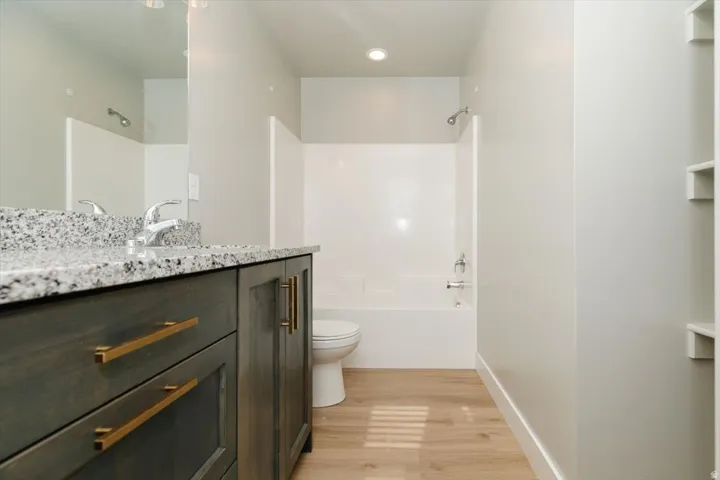 Bathroom featuring a vanity with a speckled countertop, a chrome faucet, a white tub with integrated shower, wood-finish flooring, and built-in shelving
