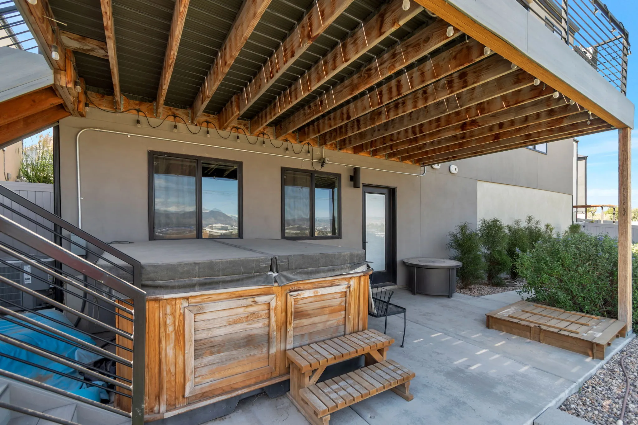 Concrete patio featuring a built-in hot tub, dark-framed sliding glass doors, a single exterior door, and a wooden deck above