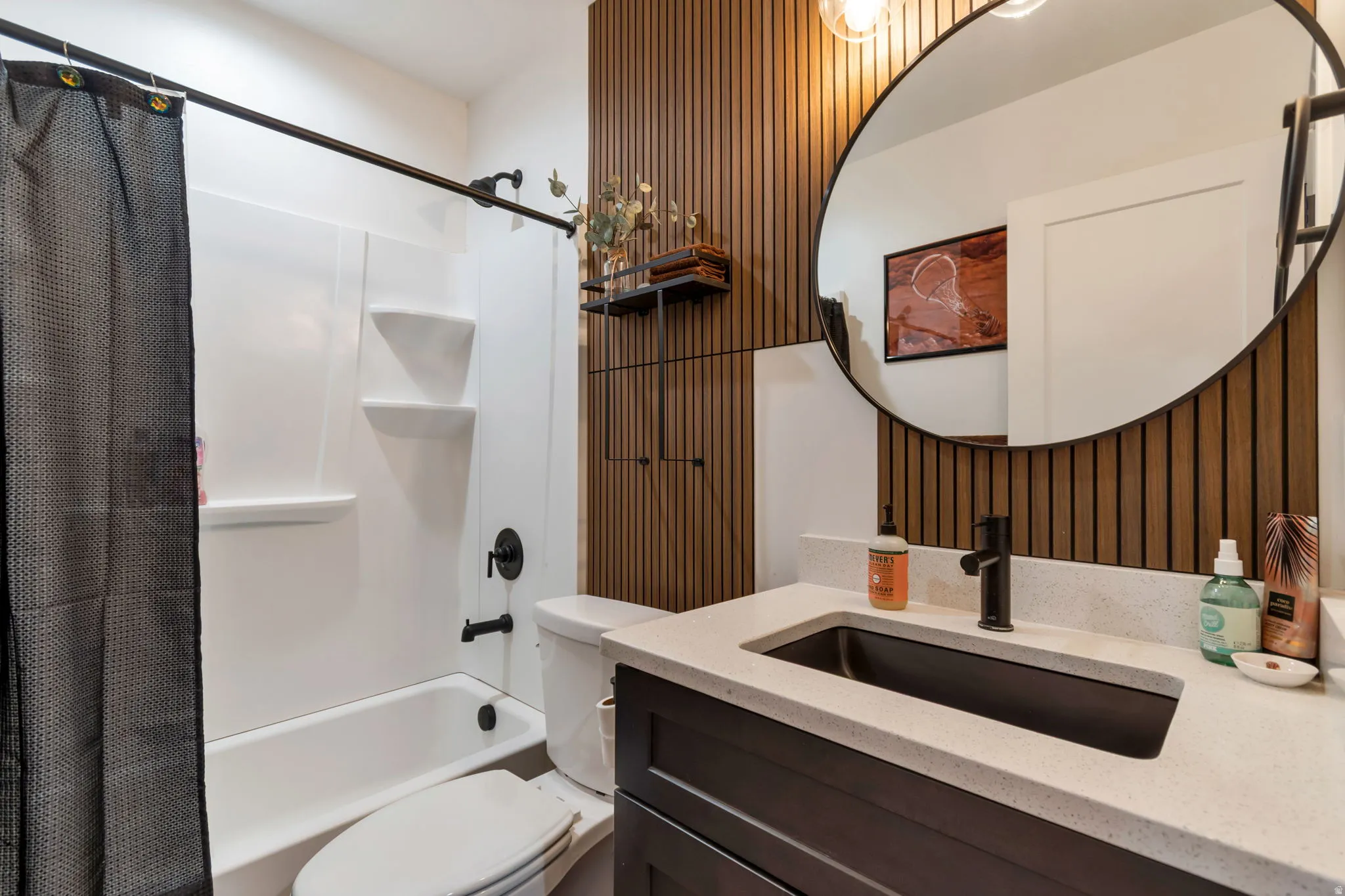White bathtub with shower, dark vanity with light countertop, black rectangular sink, black faucet, round mirror, and wood-slat accent wall