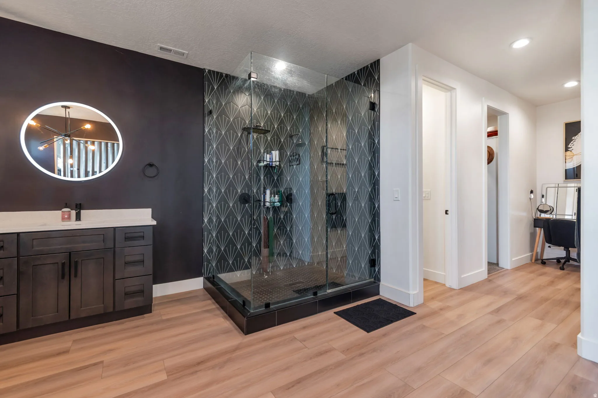 Glass-enclosed shower with patterned tile surround, wood-finish flooring, dark wood-finish vanity with light countertop, and a circular back-lit mirror