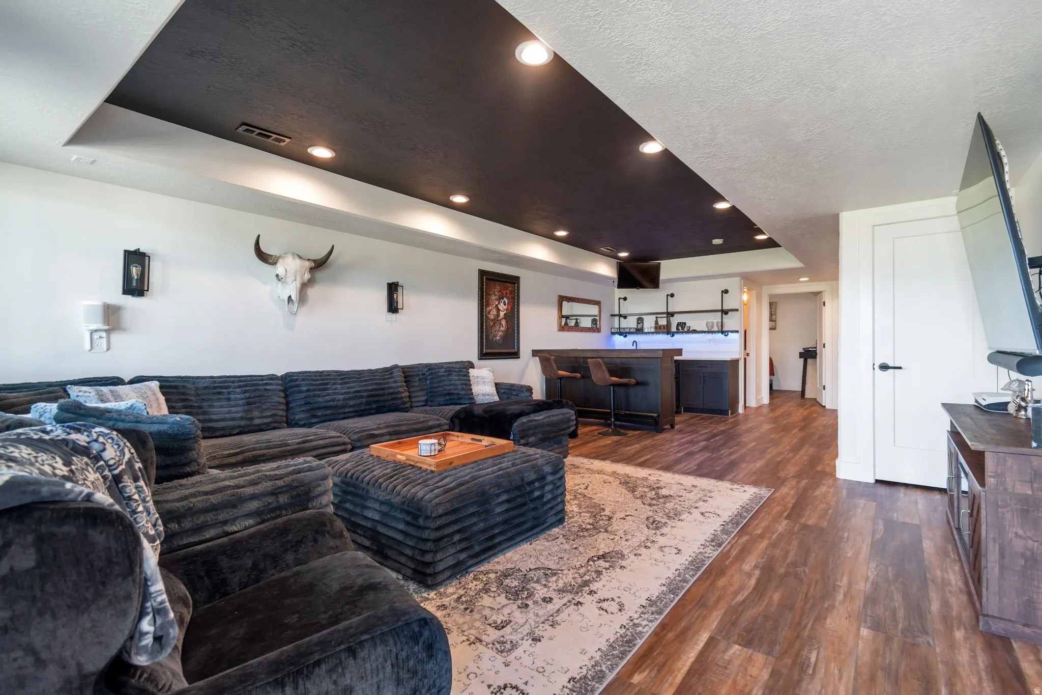 Wood-finish flooring throughout, featuring a built-in wet bar with shelving, a recessed ceiling with integrated lighting, and a solid core interior door
