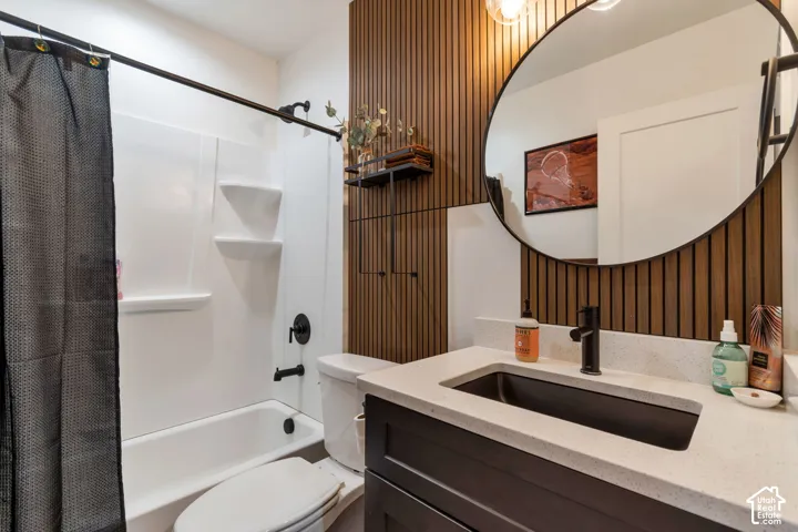 White bathtub with shower, dark vanity with light countertop, black rectangular sink, black faucet, round mirror, and wood-slat accent wall