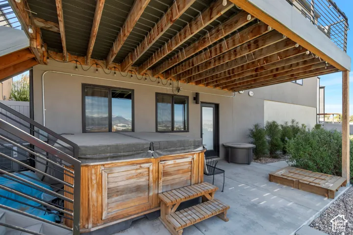 Concrete patio featuring a built-in hot tub, dark-framed sliding glass doors, a single exterior door, and a wooden deck above