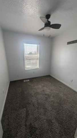 Textured gray carpeting, a window with horizontal blinds, and a ceiling fan with integrated lighting