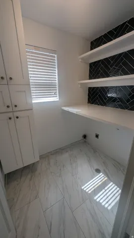 White shaker cabinetry with dark hardware, a single window with blinds, white floating shelves, black herringbone tile backsplash, and light-toned tile flooring
