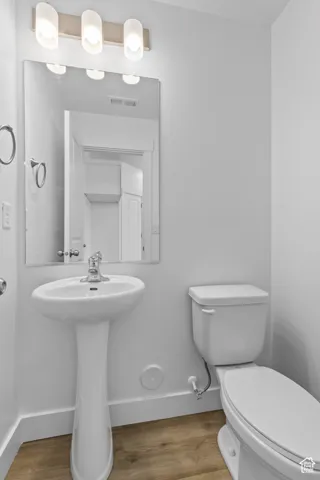 Pedestal sink with chrome faucet, wall-mounted mirror, three-light vanity fixture, white toilet, and wood-finish flooring